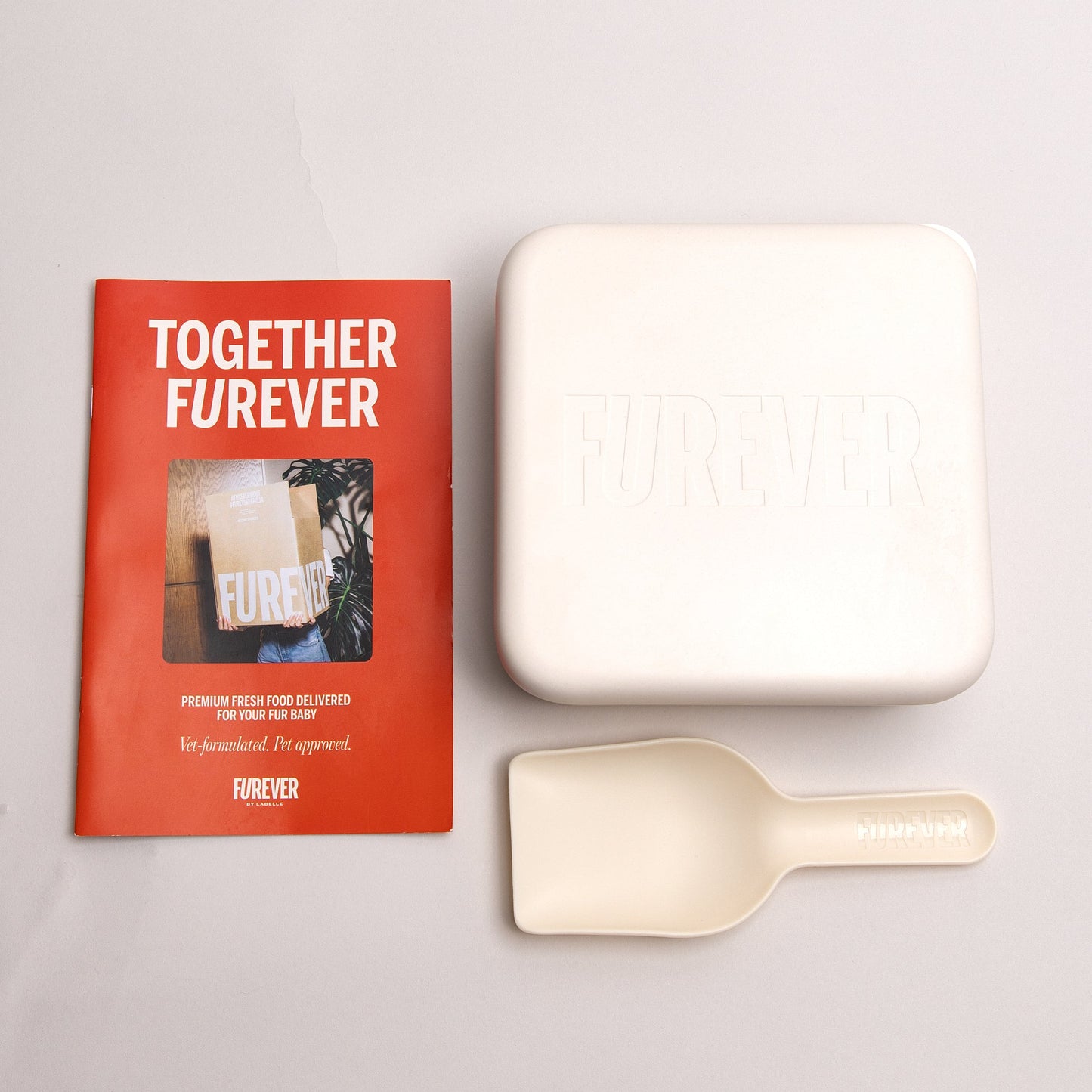 A Forever brand meal replacement set on a light gray background, featuring a red book titled 'TOGETHER FUREVER', a square off-white container embossed with 'FUREVER', and an off-white scoop with the brand name 'forever' printed on the handle.