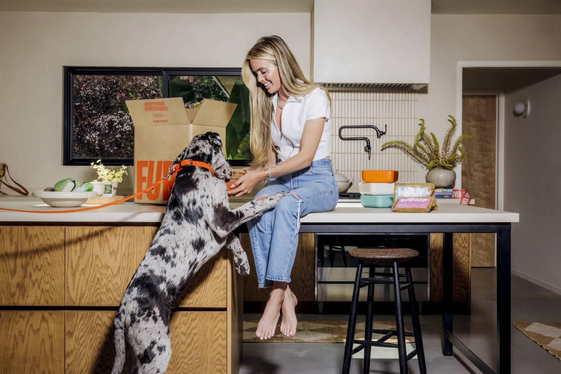 Woman sitting on a kitchen counter with a dog, surrounded by kitchen items.
